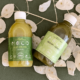 Zwei Flaschen Moco Bio-Saft, eine mit dem Etikett 'Just Celery' und die andere mit dem Etikett 'Skinny Genes', auf einem grünen Hintergrund mit weißen Dekorationsblättern.
