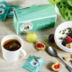 Eine Dose Thymiantee von Les 2 Marmottes auf einem Tisch, dazu eine Tasse aufgebrühten Tee, eine Schale Joghurt mit roten Früchten und Granola und frische geschnittene Feigen.