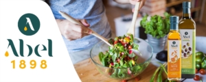 Une personne en train de préparer une salade colorée dans un grand bol en verre, mélangeant des légumes frais tels que des tomates cerises, des radis et des poivrons avec des feuilles de salade verte. Sur la gauche de l'image, le logo 'Abel 1898' apparaît, accompagné de deux bouteilles d'huile : l'une d'huile vierge de noix et l'autre d'huile d'olive extra vierge biologique. L'ambiance met en avant une cuisine saine et naturelle, avec des ingrédients frais et des huiles de qualité.